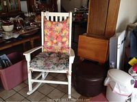 White wooden rocking chair with two floral cushions on tiled floor surrounded by various household items
