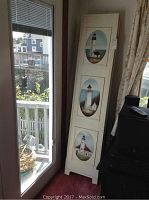 View of the three-panel folding room divider leaning next to a glass door, showing three oval lighthouse paintings on each panel.