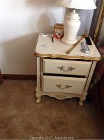 Pair of cream-colored wooden night stands with two drawers each, gold trim on the top edges. One night stand has lamp and small items on top.