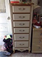 Full view of six drawer tall dresser in cream and brown finish with decorative metal handles, showing scratches and wear