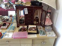 Wide shot showing wooden jewelry box with multiple drawers and mirrored panels, surrounded by plastic boxes and trays holding costume jewelry including bead necklaces, bracelets, and earrings in various colors.