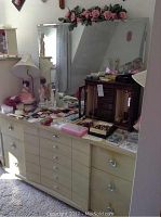 Full length frontal photo of vintage Kent-Coffey dresser with nine drawers and attached large mirror decorated with artificial pink flowers on top. Various personal items rest on the dresser surface.