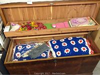 Photo showing a wooden chest containing multiple folded afghans and linens, displaying various colors and crochet patterns including floral themes.