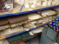 Shelf with multiple stacked unpainted ceramic figurines and shapes, showing Christmas trees and snowmen among others, stored with brown plastic containers and near blue fabric.