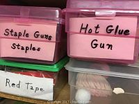 Close-up of labeled pink plastic containers with staple guns, staples, and hot glue gun. Red tape visible below.