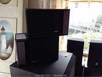 Two stacked Bose Model 141 speakers in black with rectangular shape, side and front views visible. Shown in natural light indoors near window.