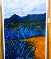 Full view of the framed oil painting depicting a desert landscape with blue vegetation and mountains under a cloudy sky.