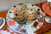 Decorative multicolored geometric patterned bowl containing three metal woven spheres in gold and bronze colors, displayed on a wooden surface.