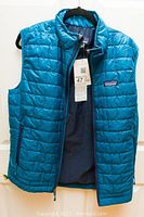 Blue men's Patagonia puffer vest hanging on a hanger, showing front and inside with visible Patagonia logo and attached tag.