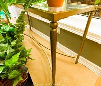 Close-up side view showing tapered gold toned metal leg and glass top edge of the sofa table beside green plants.