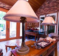 Front view of one table lamp with beige shade and ornate base on wooden surface near windowed doors.