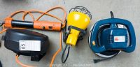 Ryobi orbital buffer in dark blue, yellow work light with cage, and orange extension cord on gray surface.