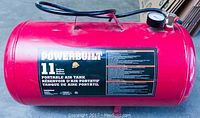 Front view of red cylindrical Power Built 11 gallon portable air tank with label, handle and connected hose and pressure gauge.