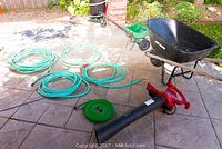 Photo showing collection of gardening tools on patio including coiled green hoses, black wheelbarrow, green spreader, and red leaf blower.