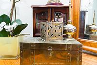 Photo showing the large brass jewelry box with a metal filigree cube and small decorative object on top, placed on a wooden dresser near a mirror and white orchid arrangement.
