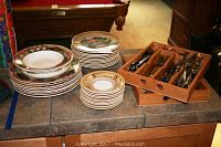 Photo of stacks of large, salad, and dessert plates with geometric patterned borders and two wooden flatware holders containing stainless steel flatware