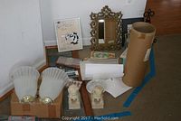 Pair of sconces with curved frosted glass and brass bases on a cardboard box, two small lion statues, framed picture, ornate mirror frame, rolled art prints and papers on floor in corner.