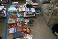 Overview photo of the book collection on the floor alongside a couch showing a variety of books in stacks and laid out flat.