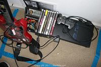 Photo showing PlayStation 2 console, two wired black controllers, red guitar controller, and a row of PlayStation 2 game cases.