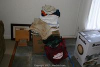 Stack of assorted linens including tablecloths and placemats on cardboard boxes in corner of room near window