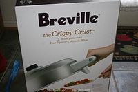 Front of the box showing the Breville brand and the product name 'the Crispy Crust 12-inch stone pizza oven' with an image of the pizza oven and a pizza being served.
