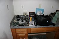 Photo of multiple electronic items arranged on a countertop including cameras, camcorder, flash, radio, and phone units.
