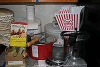 Photo showing multiple small kitchen appliances arranged on a countertop including a white electric juicer, white food dehydrator, red Manex 520 pressure cooker, silver manual popcorn popper with wooden handles, and several popcorn containers.