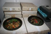 Eight boxed Russian collector plates stacked, with two plates displayed on top showing detailed scenes with colorful figures and ornate borders.