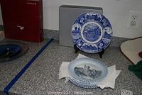 Four porcelain collector plates displayed. One plate is elevanted on a stand with Royal Copenhagen style blue scenic patterns. The other plate in front shows a black and white depiction of a castle scene with lace border.