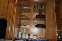 Open kitchen cabinet showing multiple shelves with ceramic kitchenware including bowls, ramekins, plates, a brown tagine pot, and utensil holders.