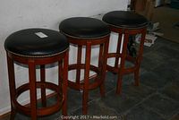 Three wooden barstools with black padded vinyl swivel seats in row against a wall on tile floor.