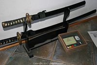 Pair of decorative swords displayed on a black stand with a small shadow box with paint set placed beside them on a tiled floor.