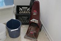 Photo showing maroon Bissell QuickSteamer PowerBrush steam cleaner on a light floor with five plastic bins behind it including a black crate and two buckets