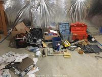 Wide shot of assorted tools, boxes, painting supplies, tools cases on a concrete floor with insulation covered walls in background.