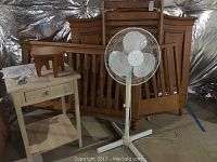 White floor fan, unfinished wooden night stand, and unassembled wooden crib parts leaning against wall.