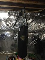 Full view of black Everlast kickboxing bag hanging from chains attached to a metal hanger bracket mounted to ceiling beams in a basement with insulation foil walls.