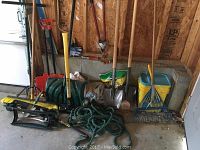 Wide shot of all items arranged against a wall: shovels, brooms, rakes, hose, storage containers, and bags.
