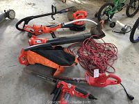 Overview photo showing multiple orange and black power tools for lawn care scattered on a concrete floor including weed trimmers, leaf blower with collection bag, and electric hedge trimmer.