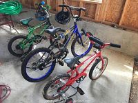 Photo of three children's bicycles in a garage on concrete floor: green bike with training wheels, blue bike without training wheels, red bike with training wheels.