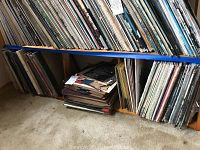 Wide view of shelves filled with numerous vinyl records stored vertically and some piled on the floor, showing a large collection.
