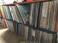 Wooden shelves densely packed with vertical stacks of vinyl records and albums in paper sleeves.