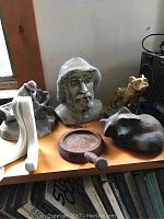 Photo showing six small statues and figures including fisherman bust with pipe, fisherman figure, cast metal raccoon, golden pug, wooden magnifying glass coaster, and wooden tusk-shaped book holder on a wood surface.