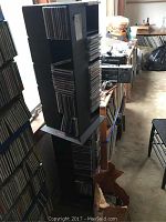 Black two-tier rotating CD tower filled with CDs seen in a cluttered room from an angle near window. Both tiers stacked vertically with CDs visible on racks and compartments.