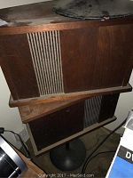 Two large vintage Bose speakers stacked on top of each other showing wood paneling and cloth grille with vertical stripes, mounted on metal pedestals.