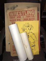 Photo showing a large vintage wrestling poster advertising a barbed wire chain match event with details in red text; also shows a yellow poster with a cartoon monkey and rolled documents.