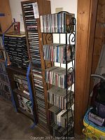 Photo showing multiple CDs arranged on a black six-tier metal CD stand with scrollwork, positioned beside a wooden wall.