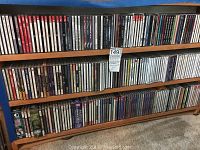 Wooden shelving with three shelves filled with over 50 assorted music CDs in cases showing spines and titles.