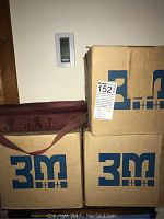 Three stacked 3M-branded cardboard boxes and a maroon canvas carrying bag underneath