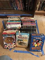 Top view of assorted cartoon DVD titles stacked on a floor surface with prominent blue tape outline, includes Betty Boop, Peanuts, Alice in Wonderland, Looney Tunes and others.