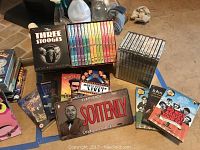Collection arranged on floor showing The Three Stooges Ultimate Collection box set, Monty Python's Flying Circus DVDs in multiple colored cases, MASH DVD set, and themed license plates and memorabilia.